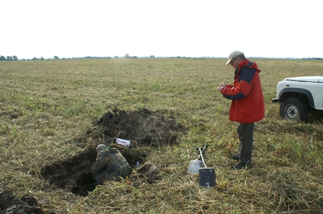 Na polu porośniętym trawą widać płytki wykop, w którym kuca jeden mężczyzna. Niedaleko wykopu stoi dr Piotrowski z notesem w rękach.