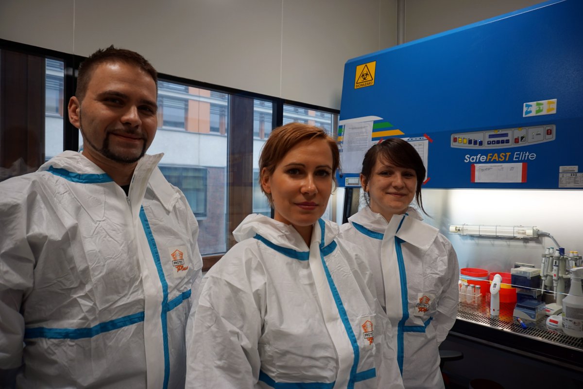 Dr hab. Krzysztof Pyrć with Dr Aleksandra Milewska and Ms Agnieszka Dąbrowska - main investigators int the project. They are standing in the laboratory on the background of research equipment, all dressed in protective suits.