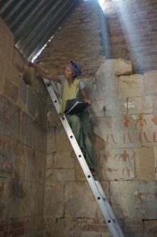 O ścianę świątyni oparta jest metalowa drabina. Na szczycie drabiny siedzi Anastasiia Stupko-Lubczyńska z otwartym zeszytem w rękach