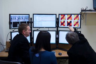 Trójka badaczy - dwóch mężczyzn i kobieta - siedzi przed sześcioma ekranami komputera. Mężczyzna siedzący po prawej wyciąga rękę w stronę jednego z ekranów, wskazując na wyświetlony tam obraz.