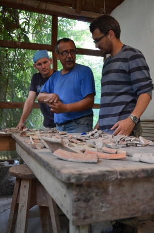 Przy drewnianym stole, na którym ułożono fragmenty ceramicznych naczyń, stoi trzech mężczyzn zajętych rozmową.